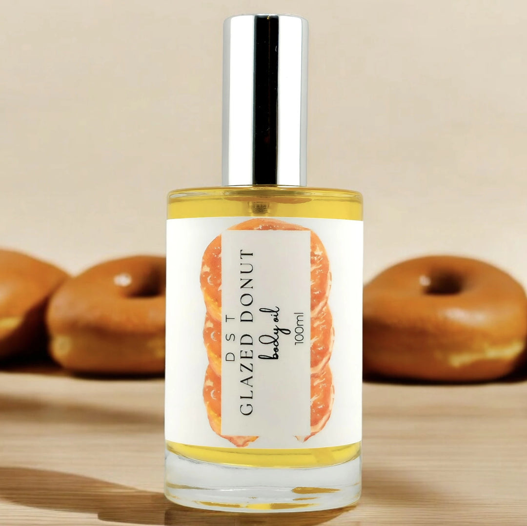 100ml bottle of Glazed Donut scented body oil sitting on a wood counter top with 3 Glazed Donuts against a tan background. 