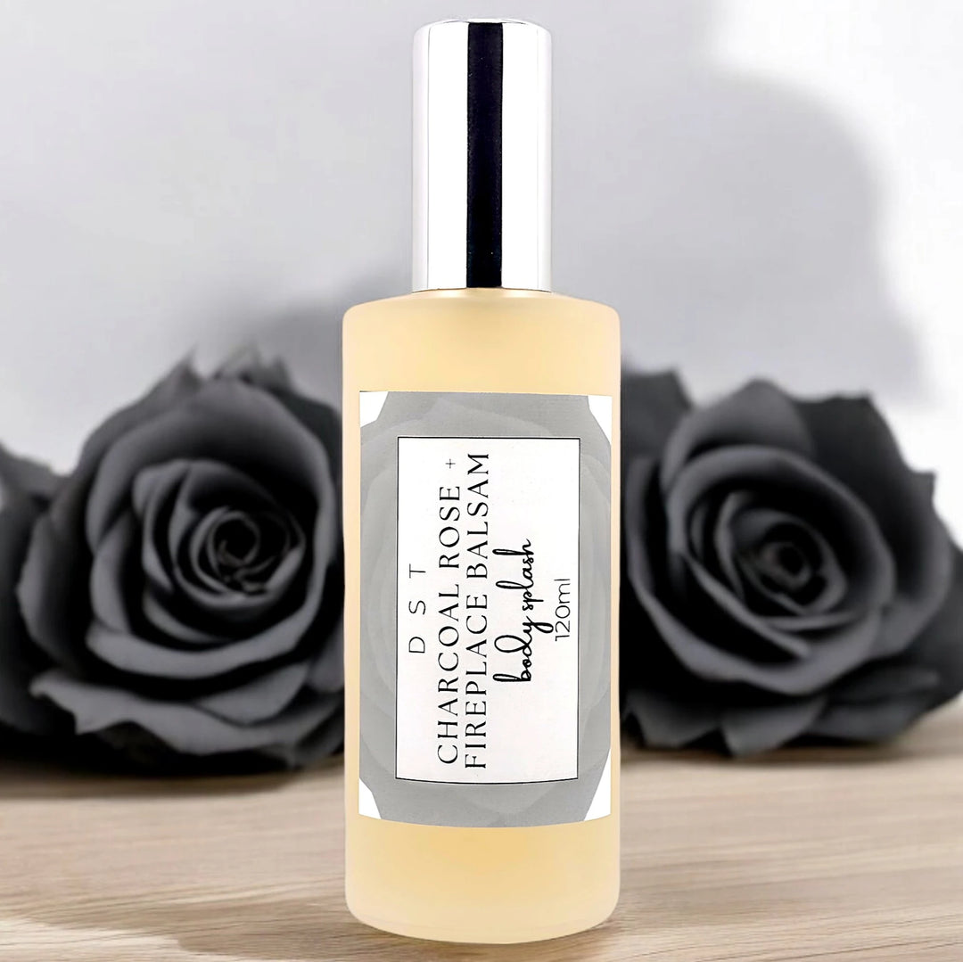 120ml bottle of Charcoal Rose and Fireplace Balsam Body splash sitting on a wood counter top with black roses against a light grey background. 