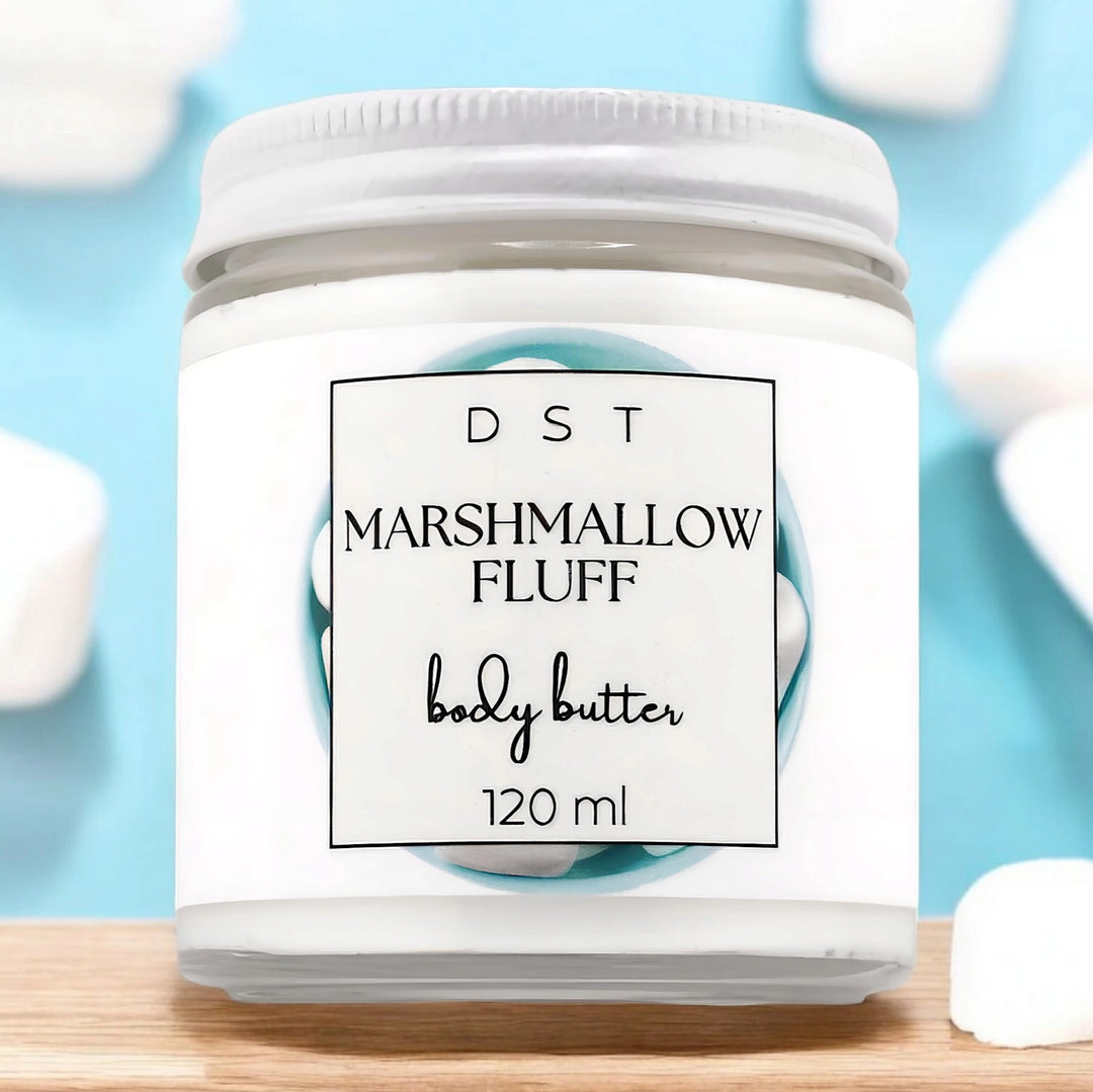 120ml jar of Marshmallow Fluff scented body butter sitting on a wood counter top with white marshmallows against a pastel blue background. 