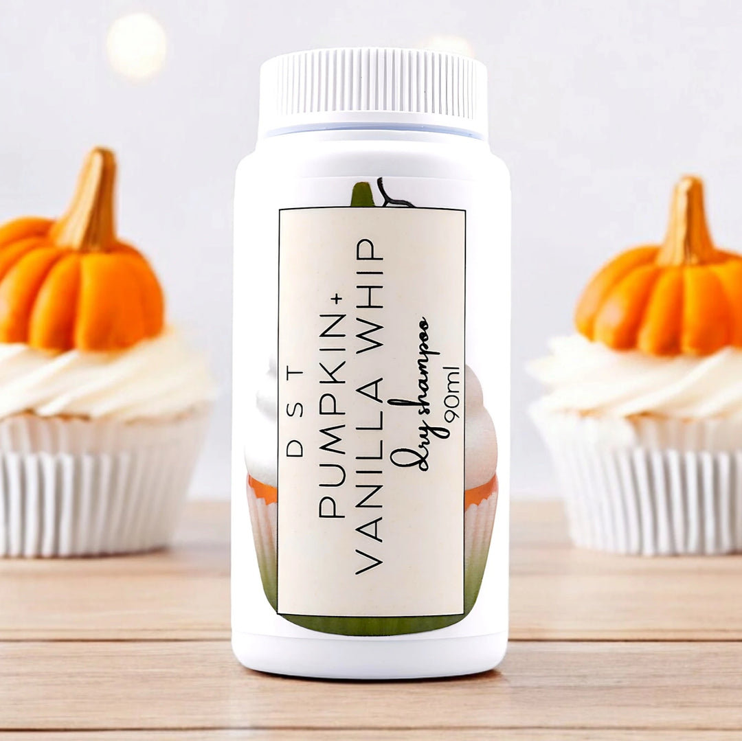 90ml bottle of Pumpkin and Vanilla Whip scented Dry Shampoo sitting on a wood counter top with 2 Vanilla cupcakes topped with fondant pumpkins against a light grey background. 