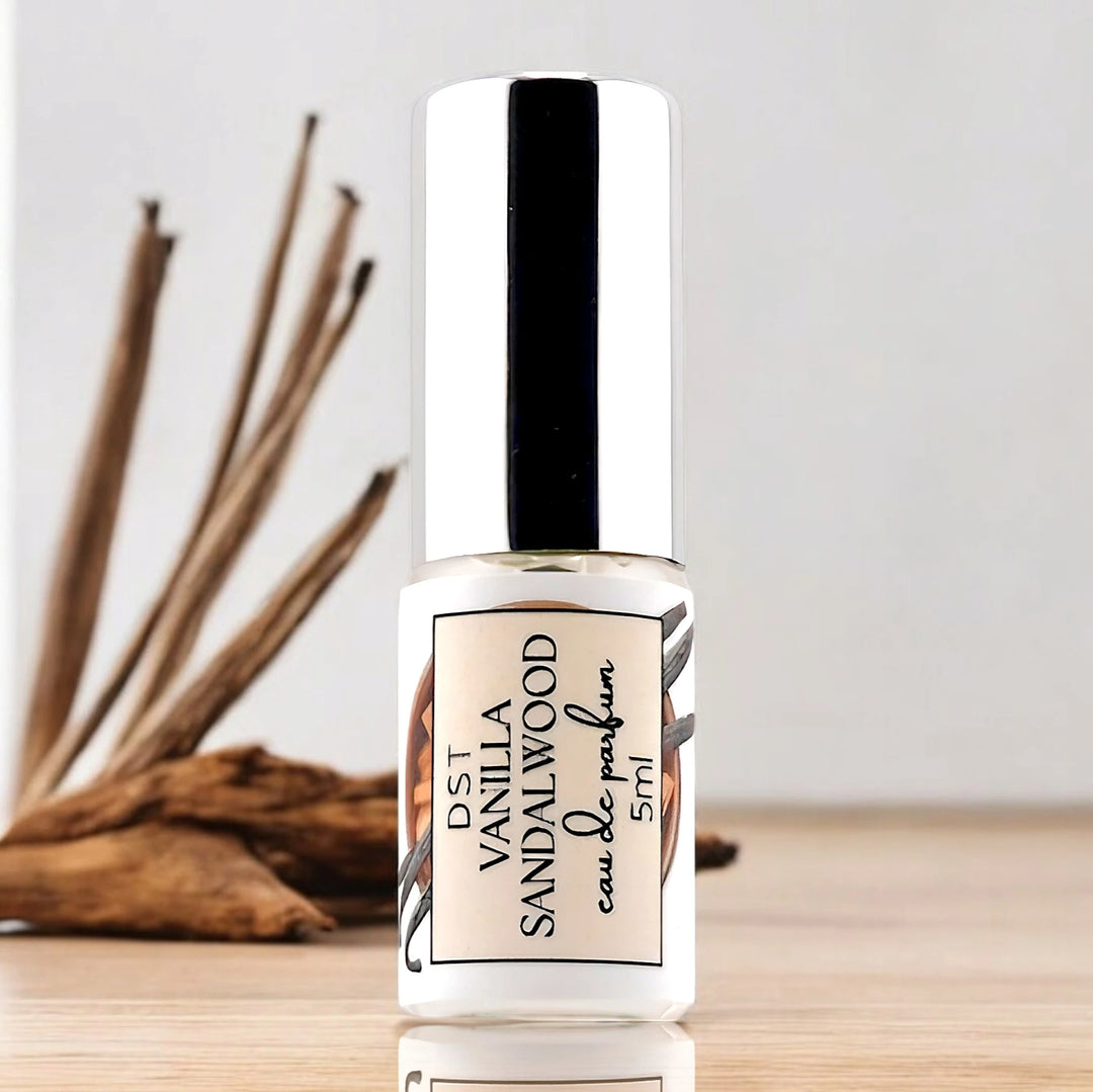 5ml bottle of Vanilla Sandalwood eau de parfum spray sitting on a wood counter top with pieces of wood and vanilla beans against a light grey background. 