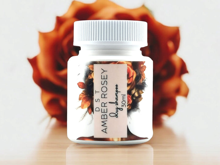 30ml bottle of Amber Rosey scented Dry Shampoo powder sitting on wood countertop with blurred orange colored rose against a white background.