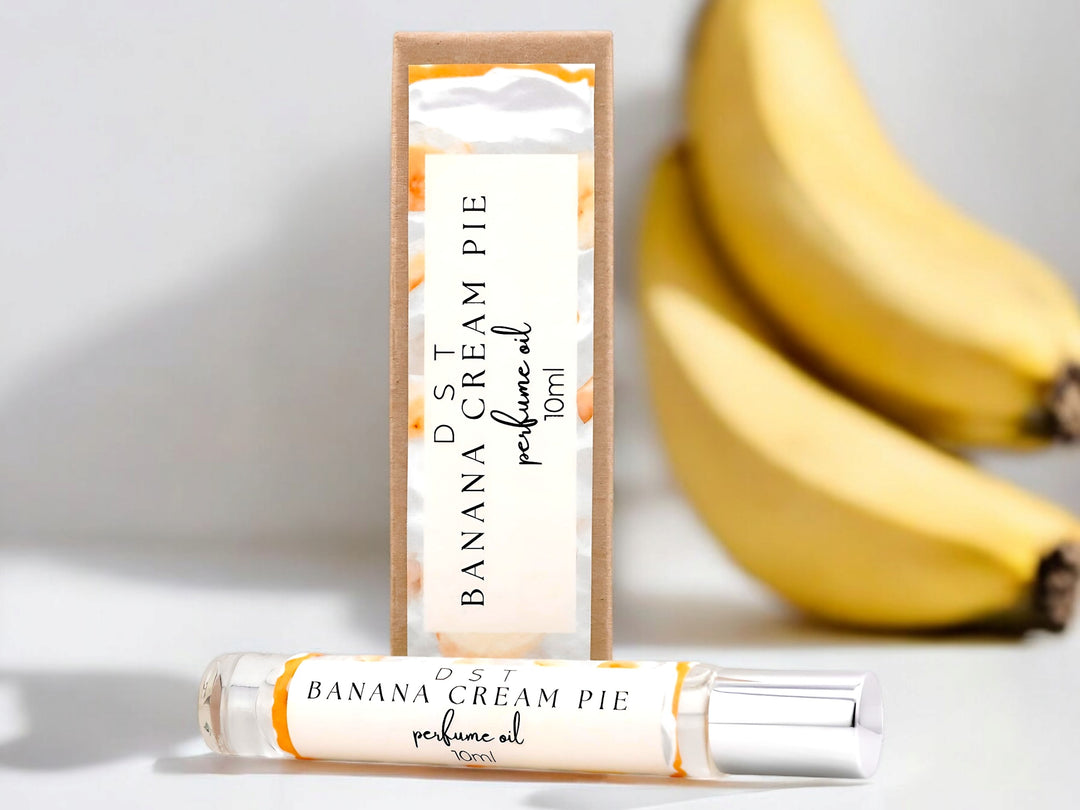 10ml bottle of Banana Cream Pie Perfume Oil and the box it is packaged in sitting on a white surface with bananas in the white shadowy background.