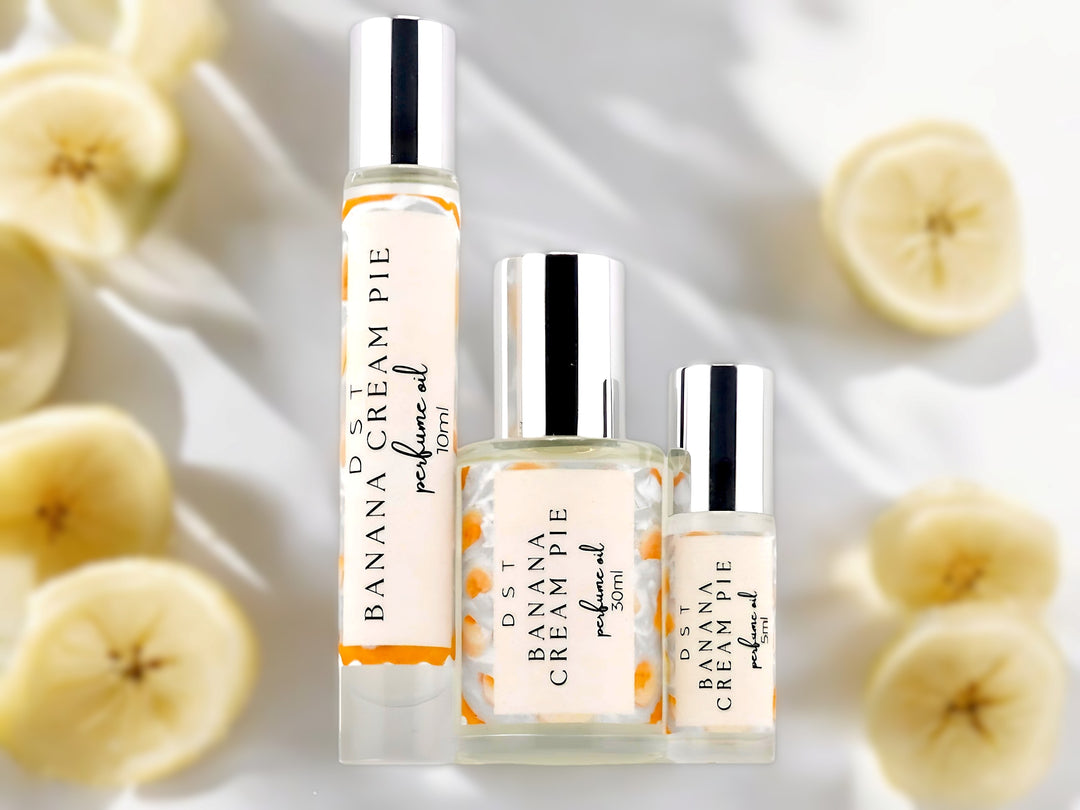 5, 10, and 30ml bottles of Banana Cream Pie Perfume Oil sitting on a glass surface with banana slices against a white shadowy background.