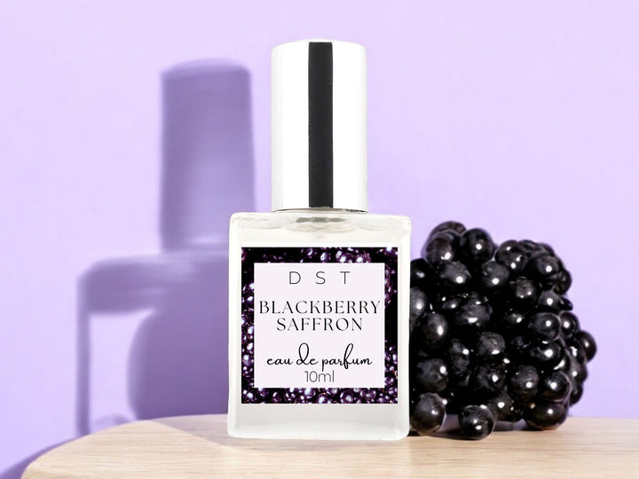 10ml bottle of Blackberry Saffron Eau de Parfum sitting on a wood countertop with a large blackberry against a pastel purple shadowy background.