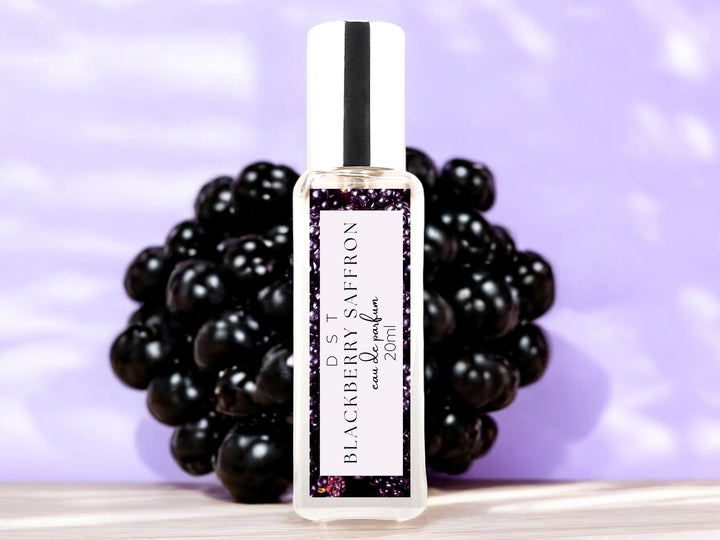 20ml bottle of Blackberry Saffron Eau de parfum spray sitting on wood countertop with large blackberry against pastel purple shadowy background.