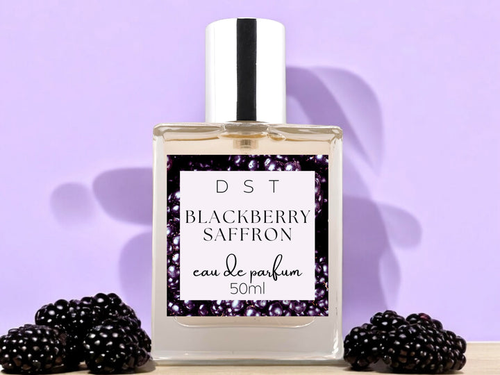 50ml bottle of Blackberry Saffron Eau de parfum spray sitting on wood countertop with blackberries against pastel purple shadowy background.