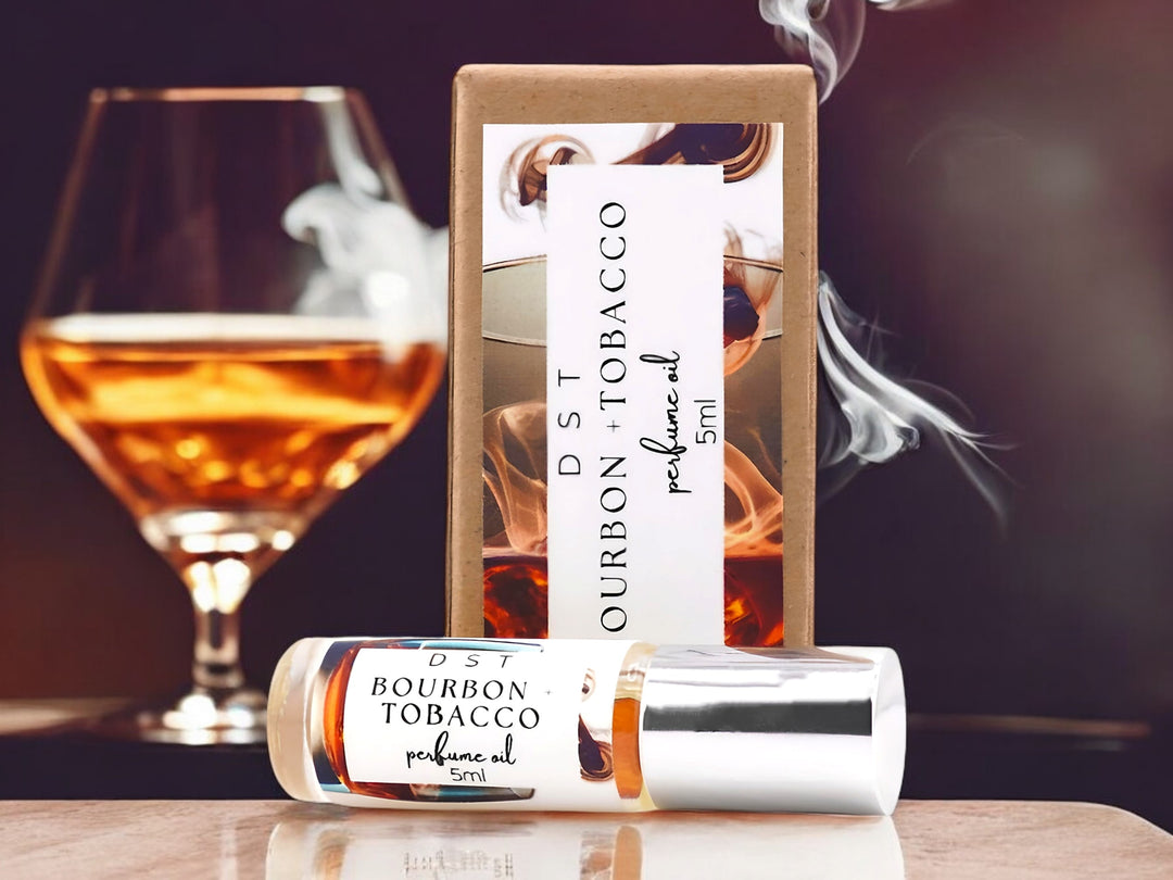 5 milliliter bottle of bourbon and tobacco perfume oil sitting on a wood countertop with a glass of bourbon and cigarette smoke behind it against a dark amber background.