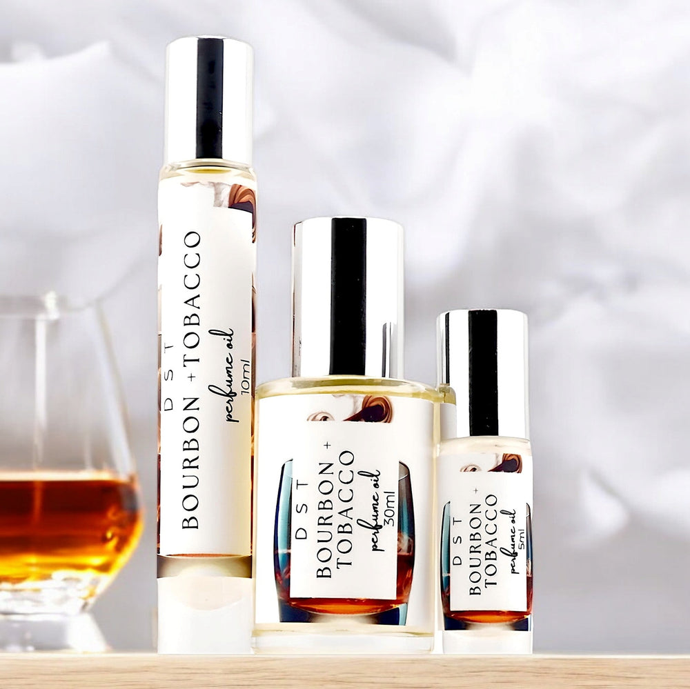 Five, 10, and 30 milliliter bottles of bourbon and tobacco perfume oil sitting on a light wood countertop with a glass of bourbon against a smoky white background.