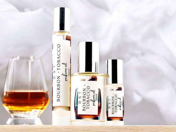 Five, 10, and 30 milliliter bottles of bourbon and tobacco perfume oil sitting on a light wood countertop with a glass of bourbon against a smoky white background.