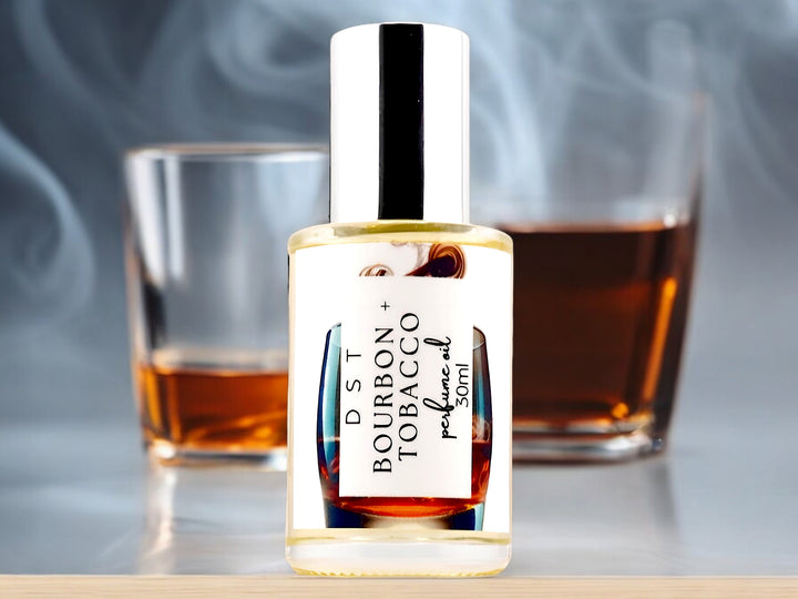 Thirty milliliter bottle of bourbon and tobacco perfume oil sitting on a light wood countertop with two glasses of Smokey Bourbon behind it against a smoky blue background.