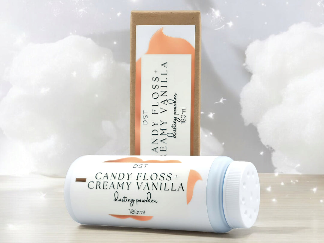 A 180ml bottle of Candy Floss and Creamy Vanilla scented body powder and the box it is packaged in sitting on wood countertop with white cotton candy and twinkling lights in the background.