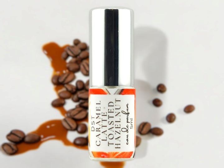 5ml bottle of Caramel Latte and Toasted Hazelnut Eau de Parfum with caramel drizzle and coffee beans against a white shadowy background.