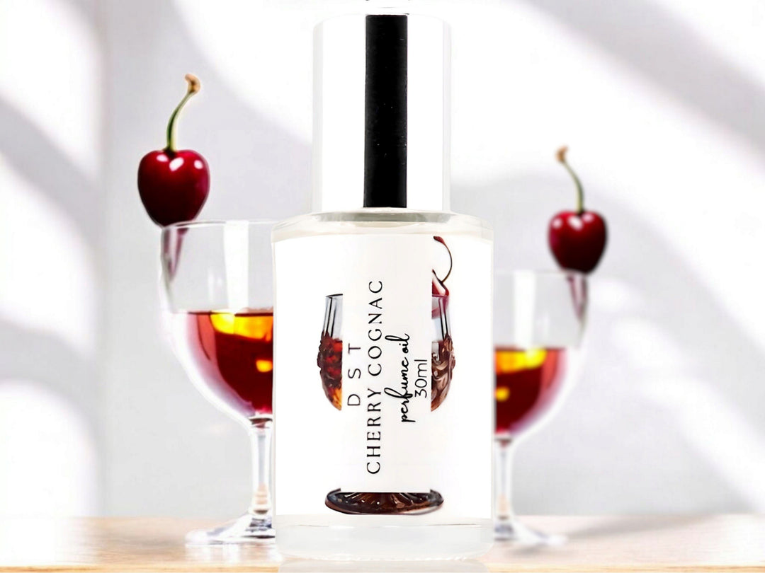 30 milliliter bottle of Cherry Cognac perfume oil sitting on a light wood countertop with two glasses of Cherry Cognac behind it against a white shadowy background.