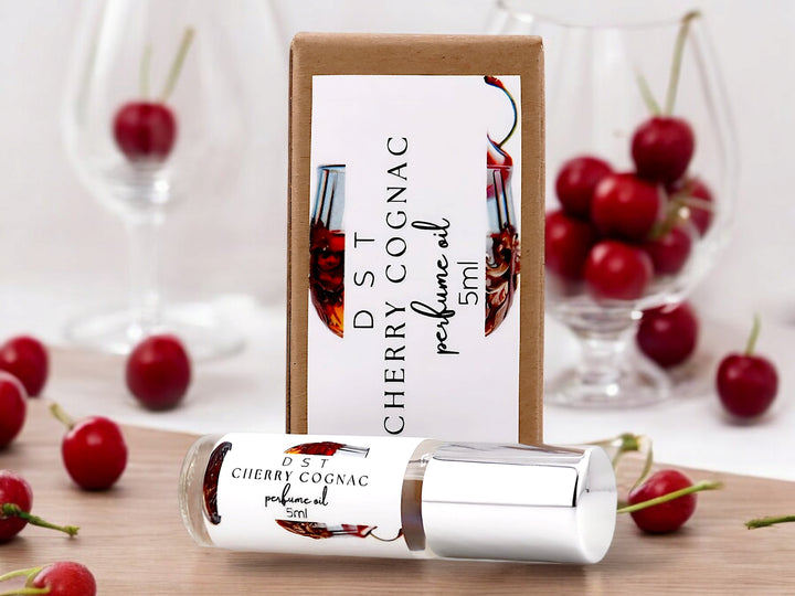 5 milliliter bottle of Cherry Cognac perfume oil and the box it is packaged in sitting on a wood countertop surrounded by scattered cherries and cognac glasses that are filled with Cherries against a blurred white background.