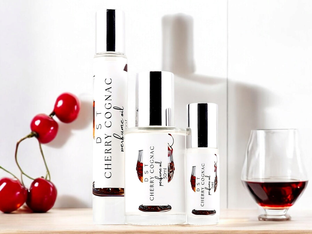 Five, 10, and 30 milliliter bottles of Cherry Cognac perfume oil sitting on a light wood countertop with a glass of cognac and scattered bright red cherries against a shadowy white background.