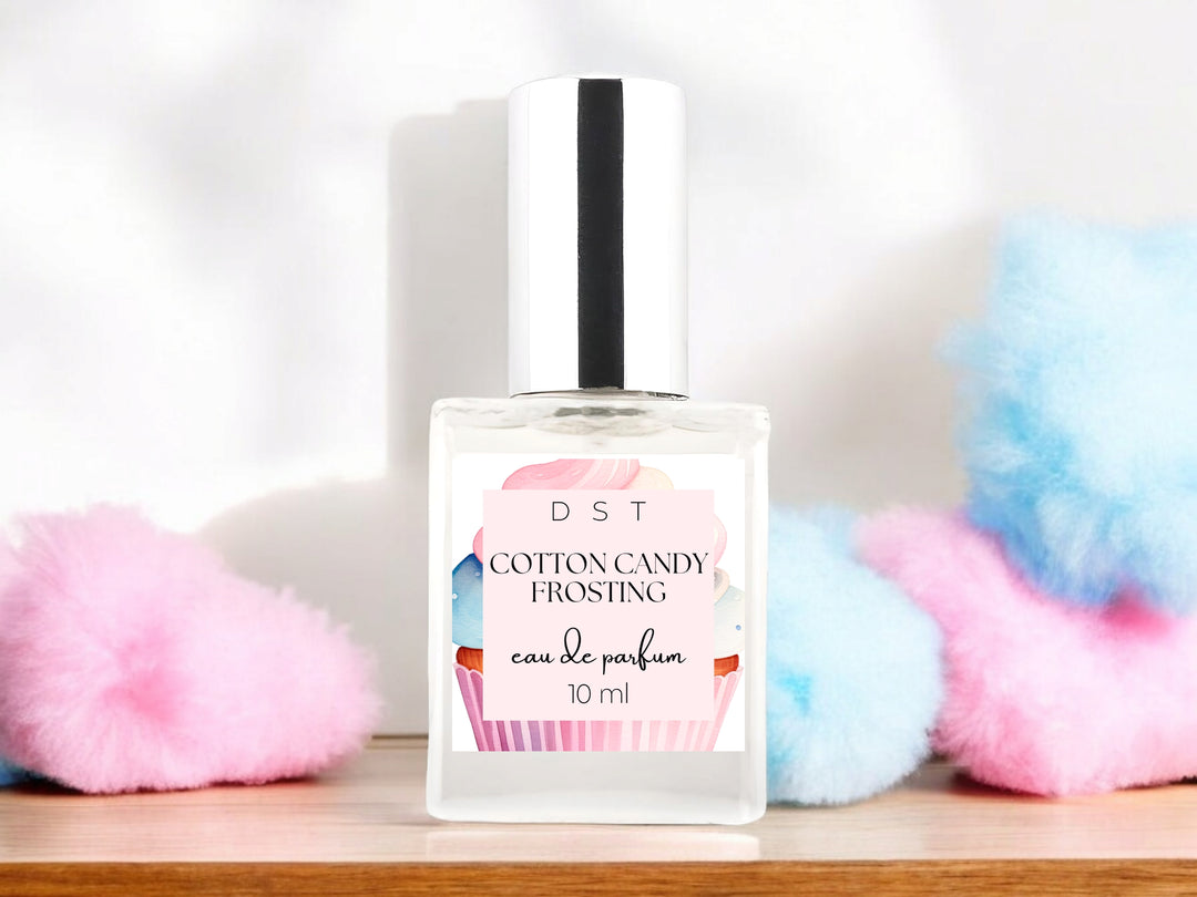 10ml bottle of Cotton Candy Frosting Eau de Parfum spray sitting on wood countertop with fluffy pink and blue cotton candy against a white shadowy background.