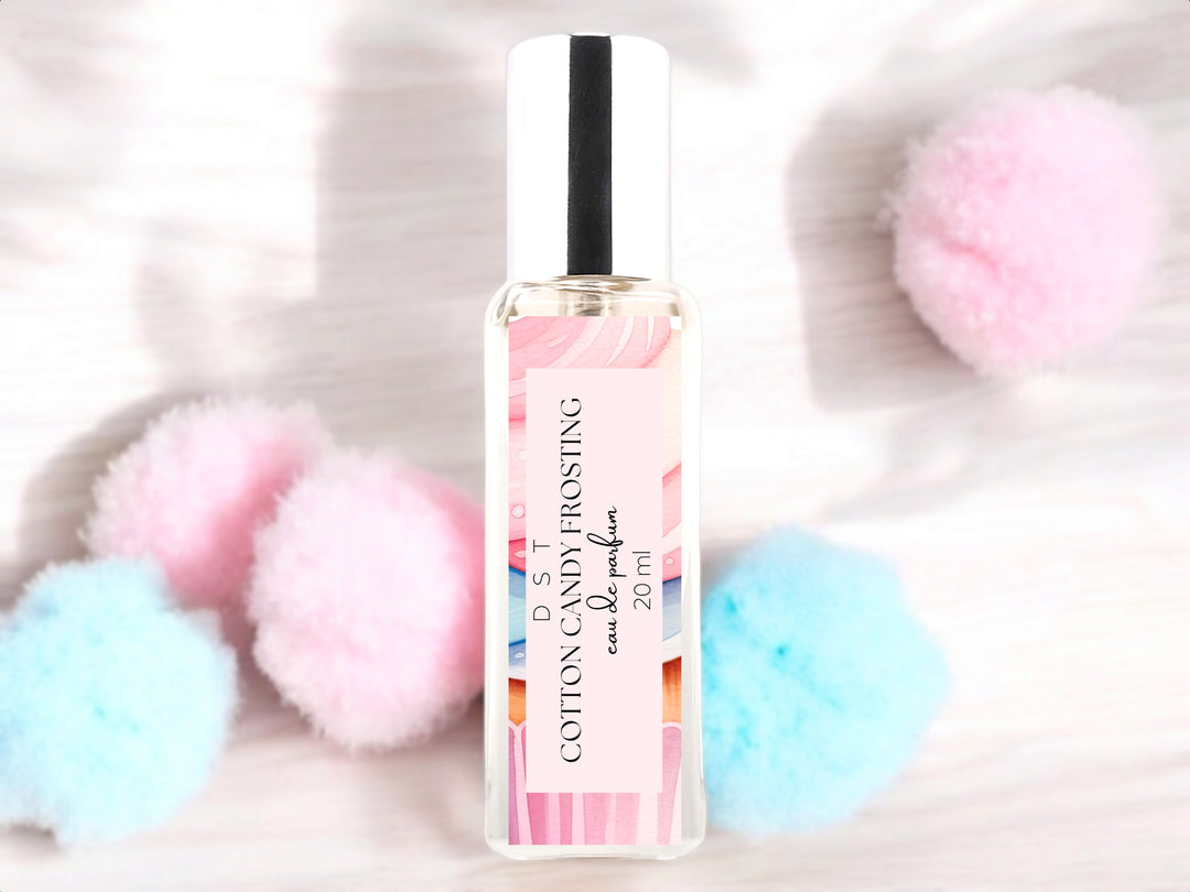 20ml bottle of Cotton Candy Frosting Eau de Parfum spray sitting on wood countertop with fluffy pink and blue cotton candy against a white shadowy background.