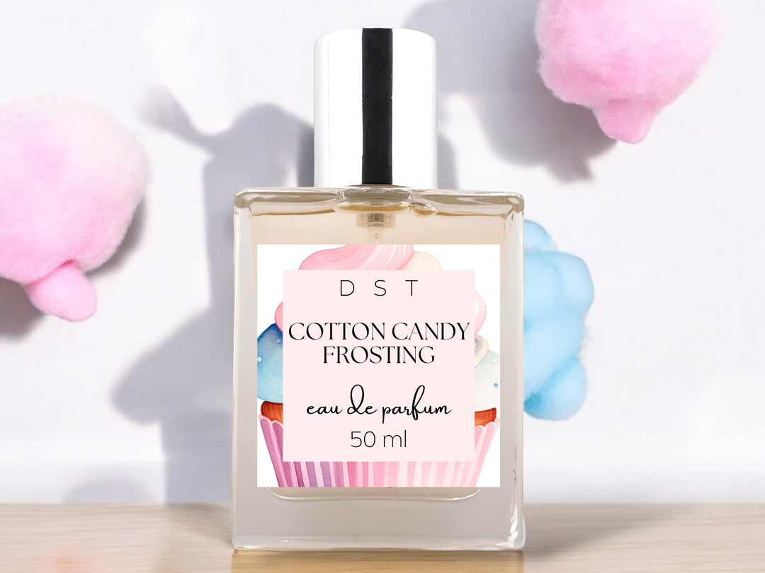 50ml bottle of Cotton Candy Frosting Eau de Parfum spray sitting on wood countertop with fluffy pink and blue cotton candy against a white shadowy background.