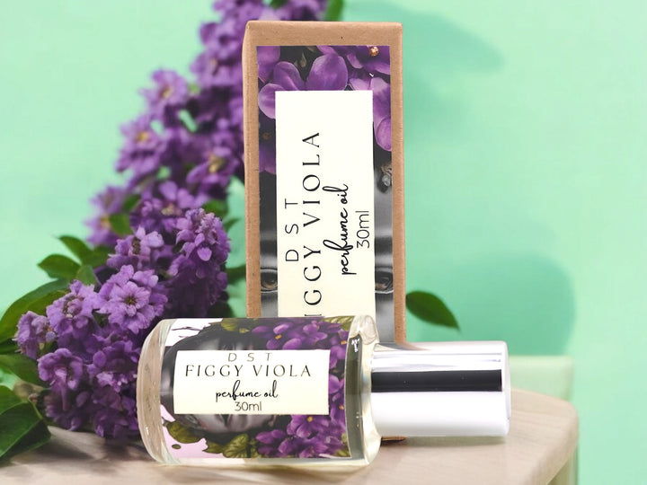 30ml Bottle of Figgy Viola perfume Oil and the box it is packaged in sitting on a wood table with violets against a pastel green background.