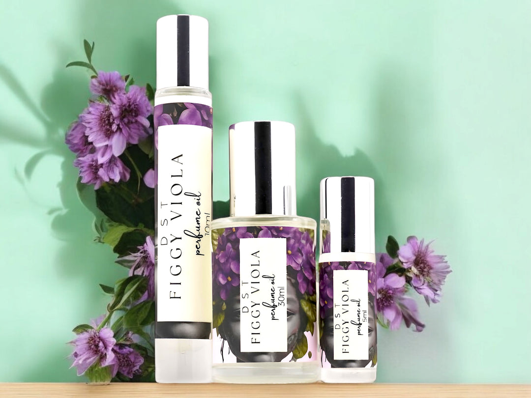 5, 10 and 30ml Bottles of Figgy Viola perfume Oil sitting on a wood table with violets against a pastel green background.