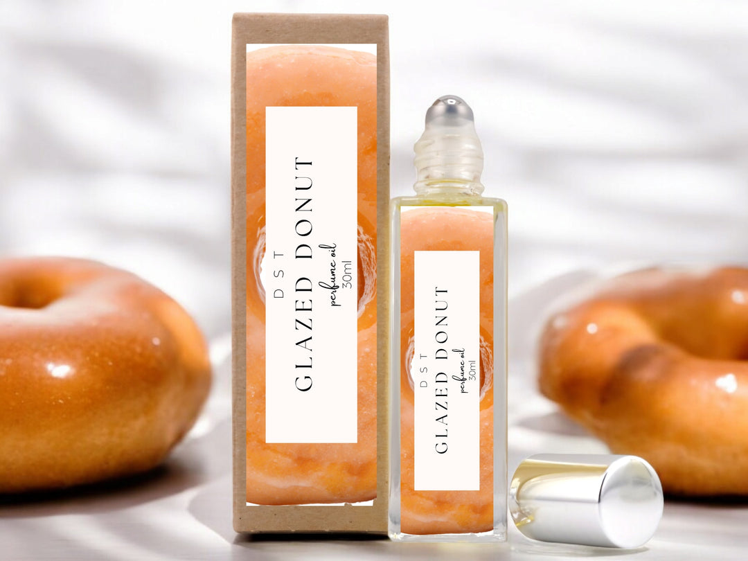 30ml bottle of Glazed Donut Perfume Oil and the box it is package in sitting on a white surface with 2 glazed donuts against a shadowy white background.