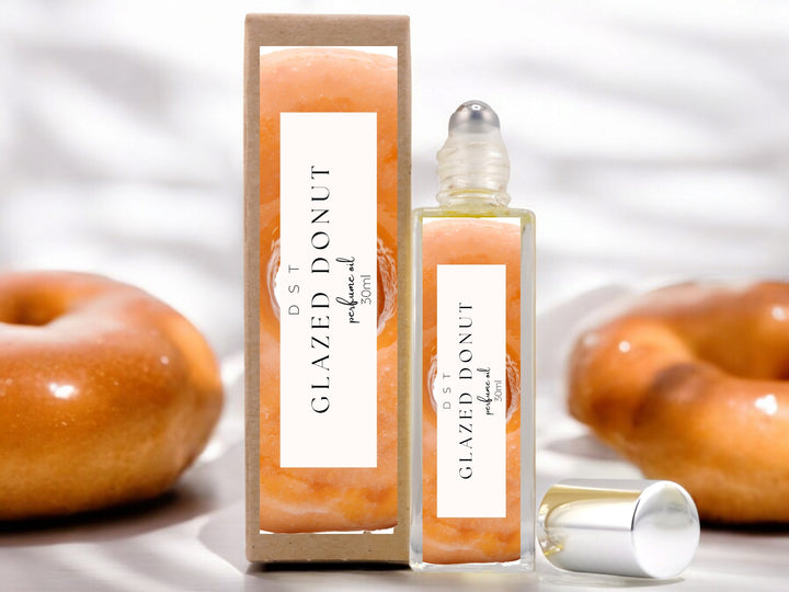 30ml bottle of Glazed Donut Perfume Oil and the box it is package in sitting on a white surface with 2 glazed donuts against a shadowy white background.