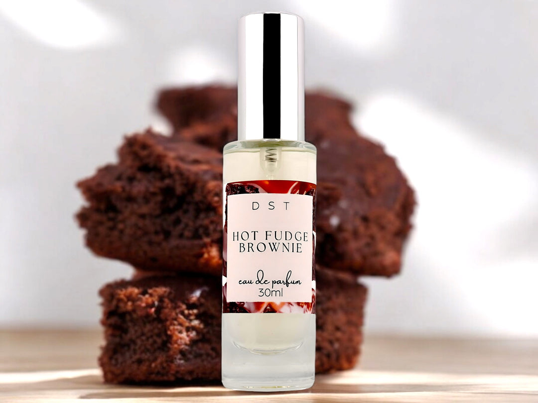 30ml bottle of Hot Fudge brownie Eau de Parfum spray sitting on a wood countertop with a stack of chocolate brownies stacked behind the perfume bottle against a white shadowy background.