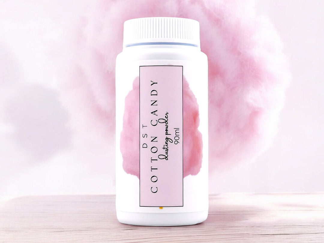 90ml bottle of Cotton Candy scented Body Powder sitting on a wood counter top with pink cotton Candy in the background.