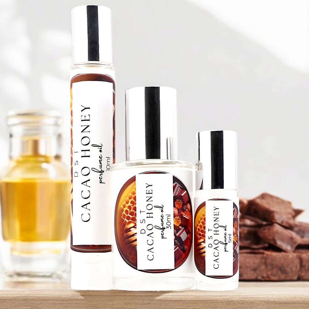 Five, ten, and thirty milliliter bottle of cacao honey perfume oil sitting on a wood countertop with chocolate chunks and a jar of honey against a shadowy white background.