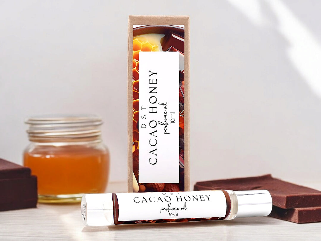 Ten milliliter bottle of cacao honey perfume oil and the box it is packaged in sitting on a wood counter top with chocolate chunks and a jar of honey against a light gray background.