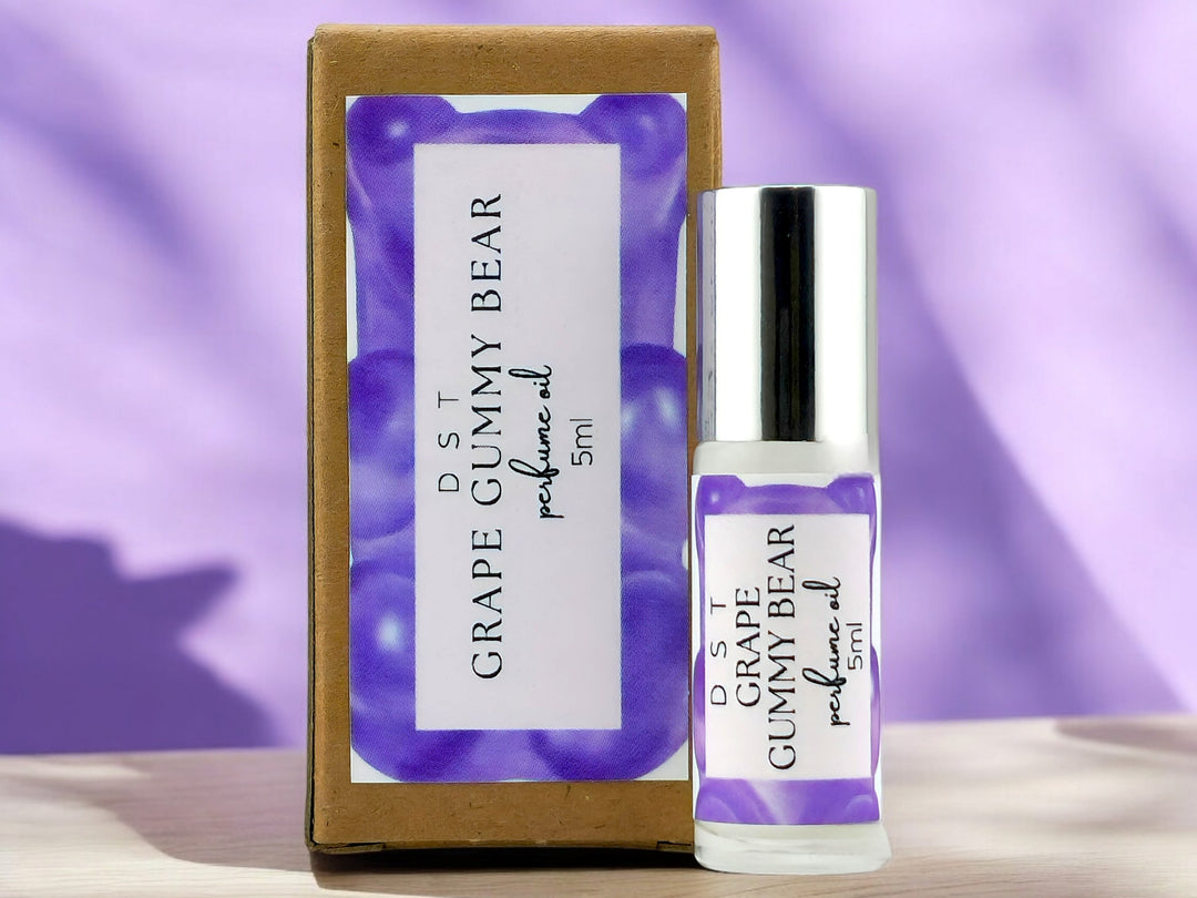 5ml bottle of Grape Gummy Bear perfume oil and the box it is packaged in sitting on wood countertop with pastel purple shadowy background.