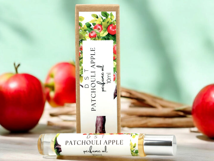 10ml bottle of Patchouli Apple perfume oil and the box it is packaged in sitting on wood countertop with red apples and cinnamon sticks in the light green background.