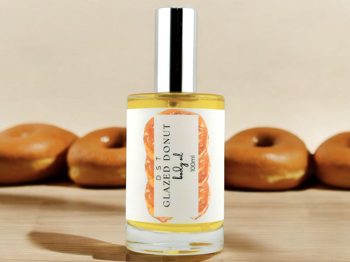 100ml bottle of Glazed Donut scented body oil sitting on wood countertop with 4 glazed donuts in the tan background.