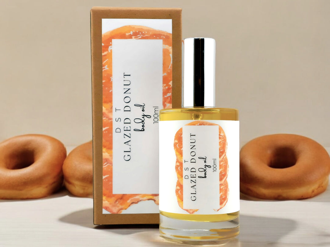 100ml bottle of Glazed Donut scented body oil and the box it is packaged in sitting on wood countertop with 3 glazed donuts in the tan background.