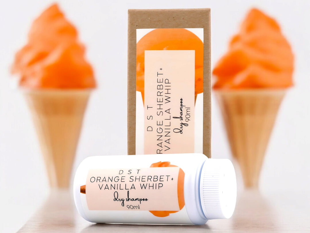 90ml bottle of Orange Sherbet and Vanilla Whip scented Dry Shampoo Powder and the box it is packaged in sitting on wood counter top with 2 sugar cones filled with orange Sherbet in the white background. 