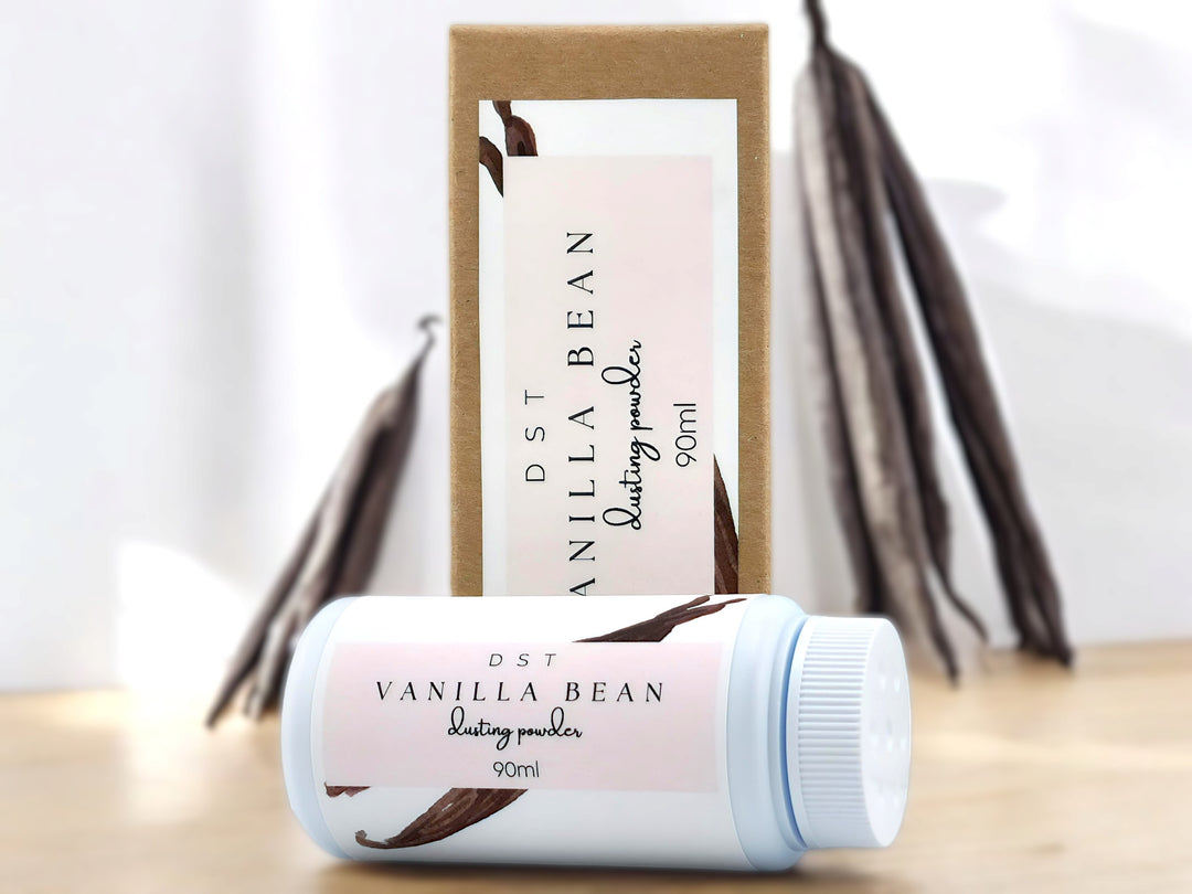 90ml bottle of Vanilla Bean powder and the box it is packaged in sitting on wood countertop with vanilla beans in the white background.