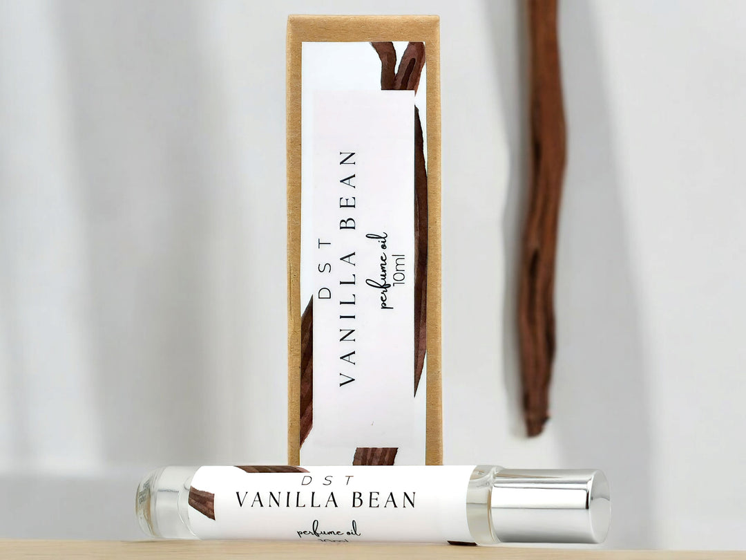 10ml bottle of Vanilla Bean perfume oil and the box it is packaged in sitting on wood countertop with white background and long vanilla bean accent.