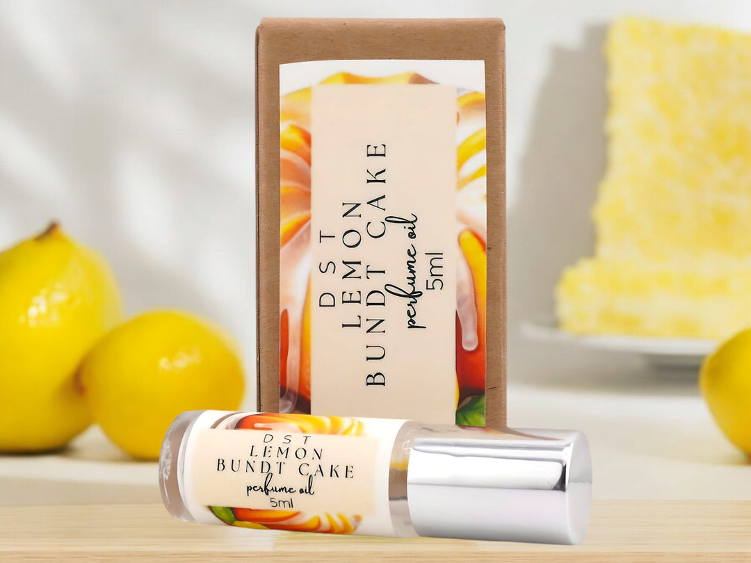 5ML bottle of lemon bundt cake perfume oil and the box it is packaged in sitting on a wood countertop with lemons and lemon cake against a white background