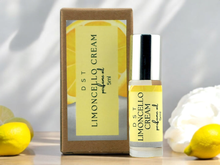 5ml bottle of Limoncello Cream perfume oil and the box it is packaged in sitting on wood countertop with lemons and whipped cream against a shadowy white background.