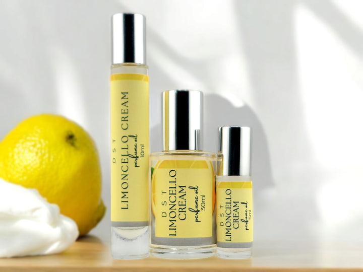 5, 10 and 30ml bottles of Limoncello Cream perfume oil sitting on wood countertop with a lemon and whipped cream against a shadowy white background.