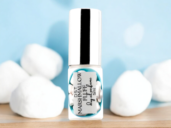 5ml bottle of Marshmallow Fluff Dry Oil spray sitting on wood countertop with puffy white marshmallows against a pastel blue background.