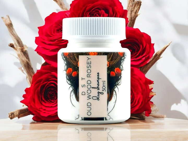 30ml bottle of Oud Wood Rosey scented Dry Shampoo powder sitting on wood countertop with red roses and wood sticks against a shadowy white background.