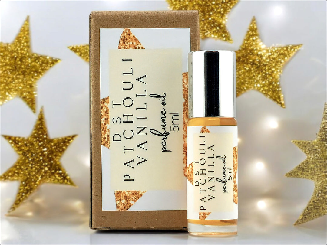 5ml bottle of Patchouli Vanilla Perfume oil and the box that it is packaged in sitting on white surface with gold glitter stars and twinkling lights against a light grey background.