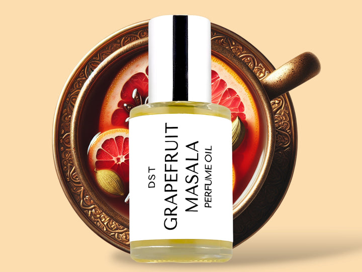 Thirty milliliter bottle of grapefruit masala perfume oil sitting in front of a large gold teacup filled with tea and topped with pink grapefruit slices and cardamom against a cream background.