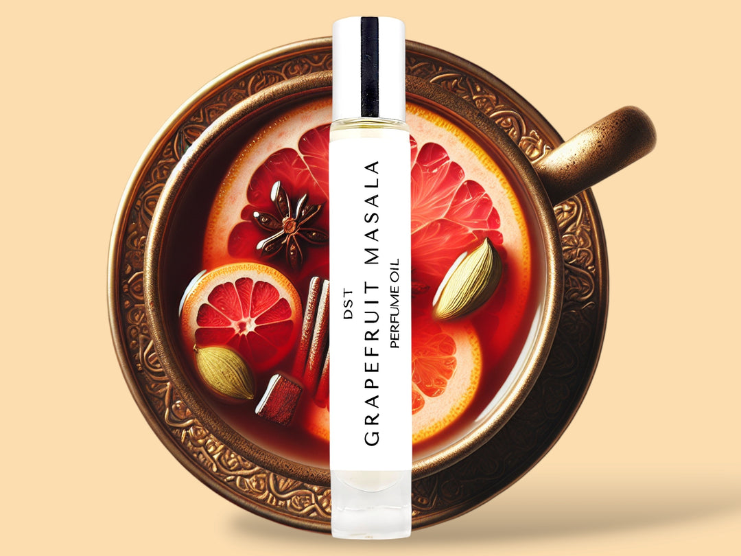 Ten milliliter bottle of grapefruit masala perfume oil sitting in front of a large gold teacup filled with tea and topped with pink grapefruit slices and cardamom against a cream background.