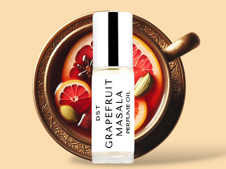 5 milliliter bottle of grapefruit masala perfume oil sitting in front of a large gold teacup filled with tea and topped with pink grapefruit slices and cardamom against a cream background.