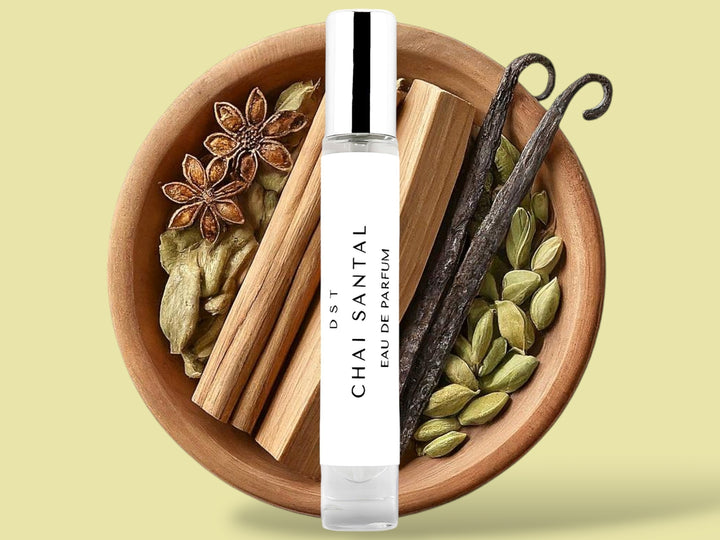 10ML bottle of Chai Santal Parfum sitting in front of a large wooded bowl filled with star anise, cardamom pods, Sandalwood, Cinnamon Sticks   and long vanilla beans, against a tan background. 