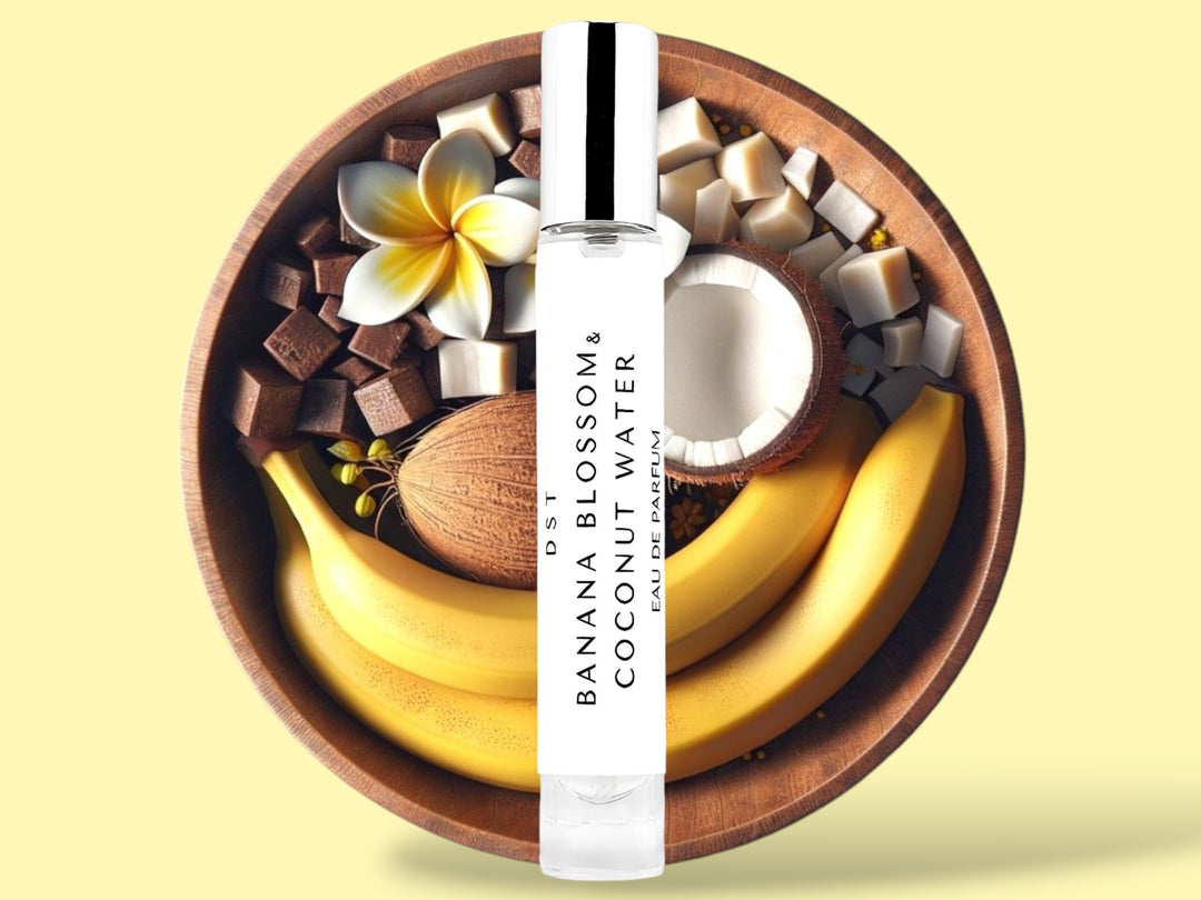 10ML bottle of Banana Blossom and Coconut Water Eau de Parfum sitting in front of a large wooden bowl filled with bananas, tiare flowers, and coconut halves, against a light yellow background. 