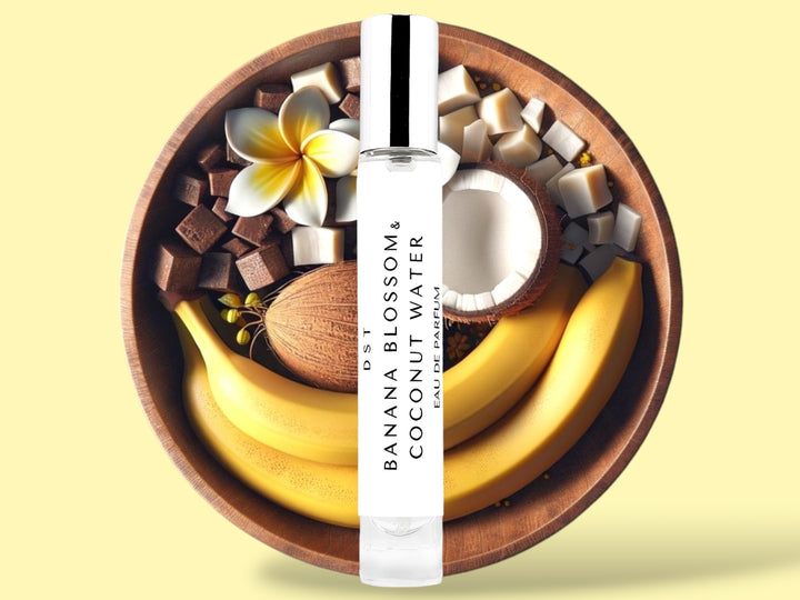 10ML bottle of Banana Blossom and Coconut Water Eau de Parfum sitting in front of a large wooden bowl filled with bananas, tiare flowers, and coconut halves, against a light yellow background. 